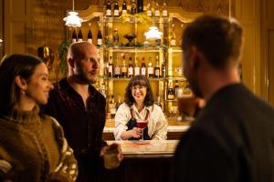 A warmly lit bar with an elegant gold-accented back wall line with liquor bottles. A smiling bartender with glasses and shoulder-length dark hair, wearing a white shirt and navy apron, serves a red cocktail in a coupe glass. In the foreground, three patrons are engaging in conversation, holding drinks. The atmosphere is inviting and sophisticated.